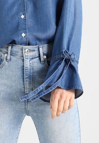 Sininen farkkupaita nappikiinnityksellä, leveillä hihoilla, joissa on solmukoristeet, yhdistettynä vaaleansinisiin farkkuihin. Sileä pinta ja rento muotoilu.