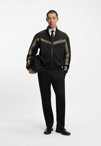 Sort atletisk zip-up jakke med brune striber, parret med sorte bukser og sko. Modellen holder et sort tøj, mens han har en hvid skjorte og slips på.