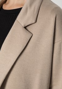 Beige textured blazer lapel over black ribbed top on person with visible neck and shoulder area.