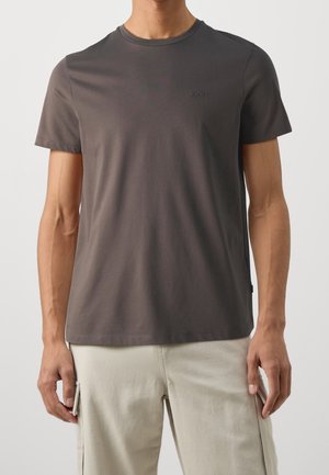Mężczyzna ubrany w ciemnoszary t-shirt z krótkim rękawem i jasnobeżowe spodnie typu cargo, stojący na tle jednolitego tła.