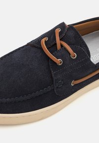 Navy suede casual shoe with tan leather laces, round eyelets, and a tan rubber sole. Features contrasting stitching and a smooth upper texture.