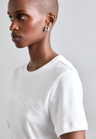 Valkoinen puuvillapaita, jossa on hienovarainen kohopainettu logo, pyöreä pääntie ja lyhyet hihat. Mallilla on päällään hopeiset rengaskorvakorut.