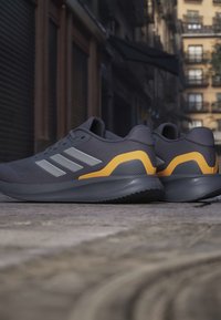 Chaussures de sport gris foncé avec des accents jaunes au talon et des bandes blanches sur les côtés, reposant sur une rue pavée dans un environnement urbain.