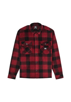 Jeune personne aux cheveux ondulés se tenant face à l'avant, vêtue d'une chemise à carreaux rouge et noire, d'un pantalon noir et de baskets noires, sur un fond uni.