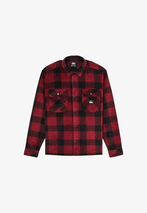 Jeune personne aux cheveux ondulés se tenant face à l'avant, vêtue d'une chemise à carreaux rouge et noire, d'un pantalon noir et de baskets noires, sur un fond uni.