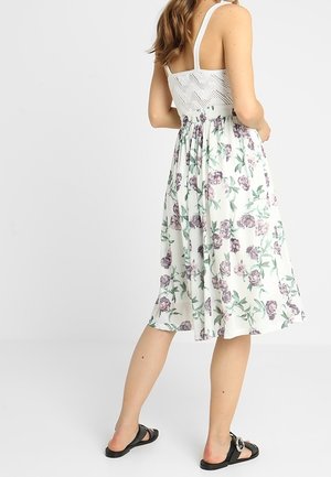 Frau trägt ein weißes ärmelloses Kleid mit lila Blumenmuster und schwarzen Sandalen, steht mit leicht gedrehtem Rücken.