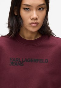 Jonge vrouw met donker haar, gekleed in een bordeauxrode sweatshirt met 'KARL LAGERFELD JEANS' in het zwart op de borst gedrukt.