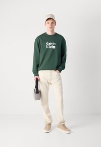 Zelená mikina s bielym logom "Calvin Klein", krémové nohavice, béžové tenisky a svetlosivá čiapka, drží šedú crossbody tašku.