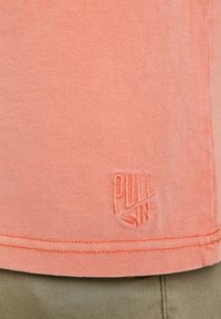 T-shirt en coton couleur pêche avec texture côtelée, arborant un logo brodé "PULL IN" à l'ourlet. Coupe droite avec coutures finies.