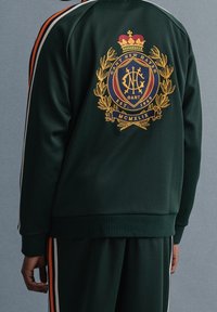 Chaqueta deportiva verde con dobladillo acanalado, que presenta un escudo bordado en dorado en la parte posterior y rayas laterales en naranja y blanco.