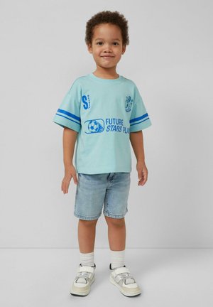 Glimlachende jonge jongen met krullend haar, gekleed in een lichtblauw voetbal-T-shirt met de tekst "Future Stars Play", spijkerbroekshort, witte sokken en sneakers.