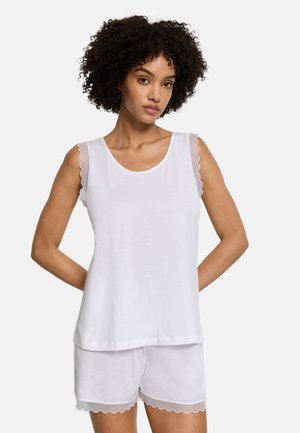 Frau trägt weißes ärmelloses Oberteil und passende Shorts mit gewelltem Spitzenbesatz, steht mit den Händen hinter dem Rücken.