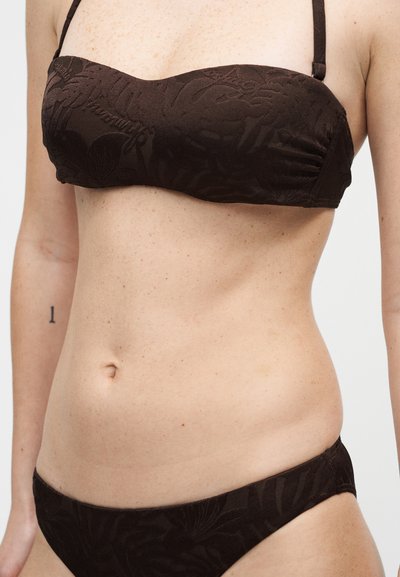 Γυναίκα που φοράει σκούρο καφέ υφασμάτινο σετ μπικίνι με λεπτές τιράντες, εμφανίζοντας τον κορμό και μερικά χέρια με ένα μικρό τατουάζ "1" στο αριστερό χέρι.