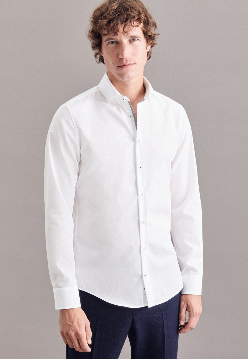 Camicia bianca a maniche lunghe in tessuto morbido, con chiusura a bottoni e colletto rigido, abbinata a pantaloni scuri.