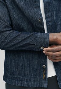 Nærbillede af en persons hånd, der justerer knappen på en mørk stripet denimjakke med en hvid skjorte under og et synligt mærke.