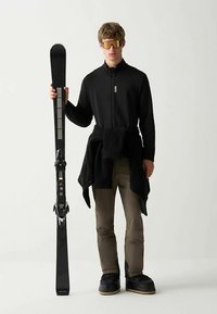 Fekete cipzáras dzseki fekete polárfelső fölött, színes barna nadrág és fekete bakancs. A modell fekete síléccel pózol. A megjelenést tükörhatású napszemüveg teszi teljessé.