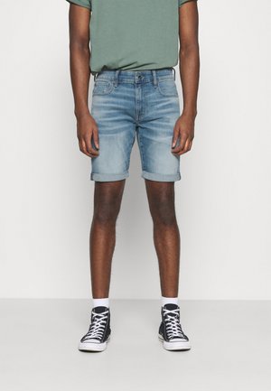 Jasnoniebieskie szorty z denimu z podwiniętymi mankietami, w połączeniu z zielonym t-shirtem z krótkim rękawem oraz czarnymi wysokimi trampkami z białymi podeszwami.