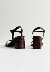 Pareja de sandalias de tacón marrón oscuro con correas en el tobillo y tacones de bloque, exhibidas sobre una superficie blanca con un fondo neutro.