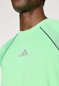 Light green short-sleeve athletic shirt with black accents and a gray logo. Features a round neckline and textured mesh panels on shoulders.