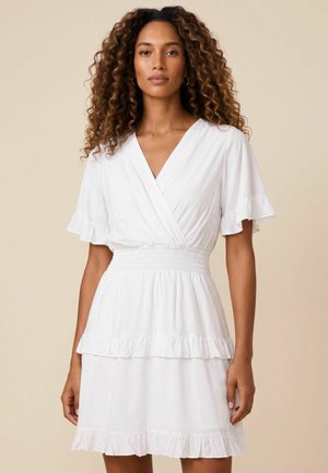 Femme aux cheveux bouclés portant une robe blanche à manches courtes avec des bords volantés et une taille froncée, debout devant un fond uni.