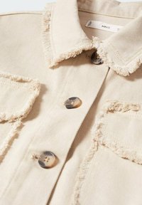 Beige denimjakke med flossede kanter, der har en spids krave, to brystlommer og runde knapper med et marmor-mønster.