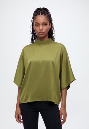 Femme aux cheveux tressés portant des boucles d’oreilles créoles en or, un haut ample à manches courtes vert olive avec un col montant et un pantalon noir.