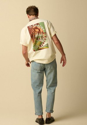 Man wearing light yellow T-shirt with colorful botanical and architectural print and text, paired with light blue jeans and black shoes.