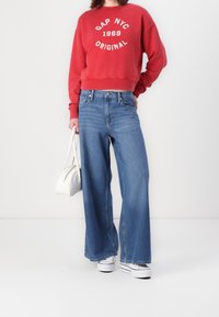 Sudadera roja corta con estampado blanco, jeans de pierna ancha azules y zapatillas blancas. El conjunto incluye un pequeño bolso blanco.