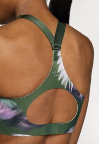 Zelená športová podprsenka s výrezom na zadnej strane, hodvábnymi ramienkami a kvetinovým vzorom v odtieňoch fialovej a zelenej.