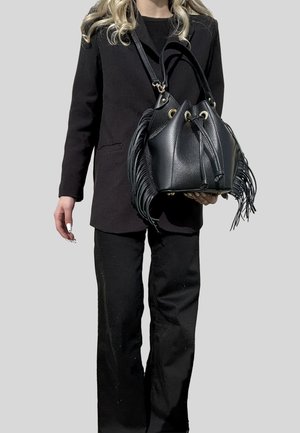 Donna in abito nero che tiene una borsa a secchiello di pelle nera con dettagli a frange e chiusura con coulisse su sfondo neutro.
