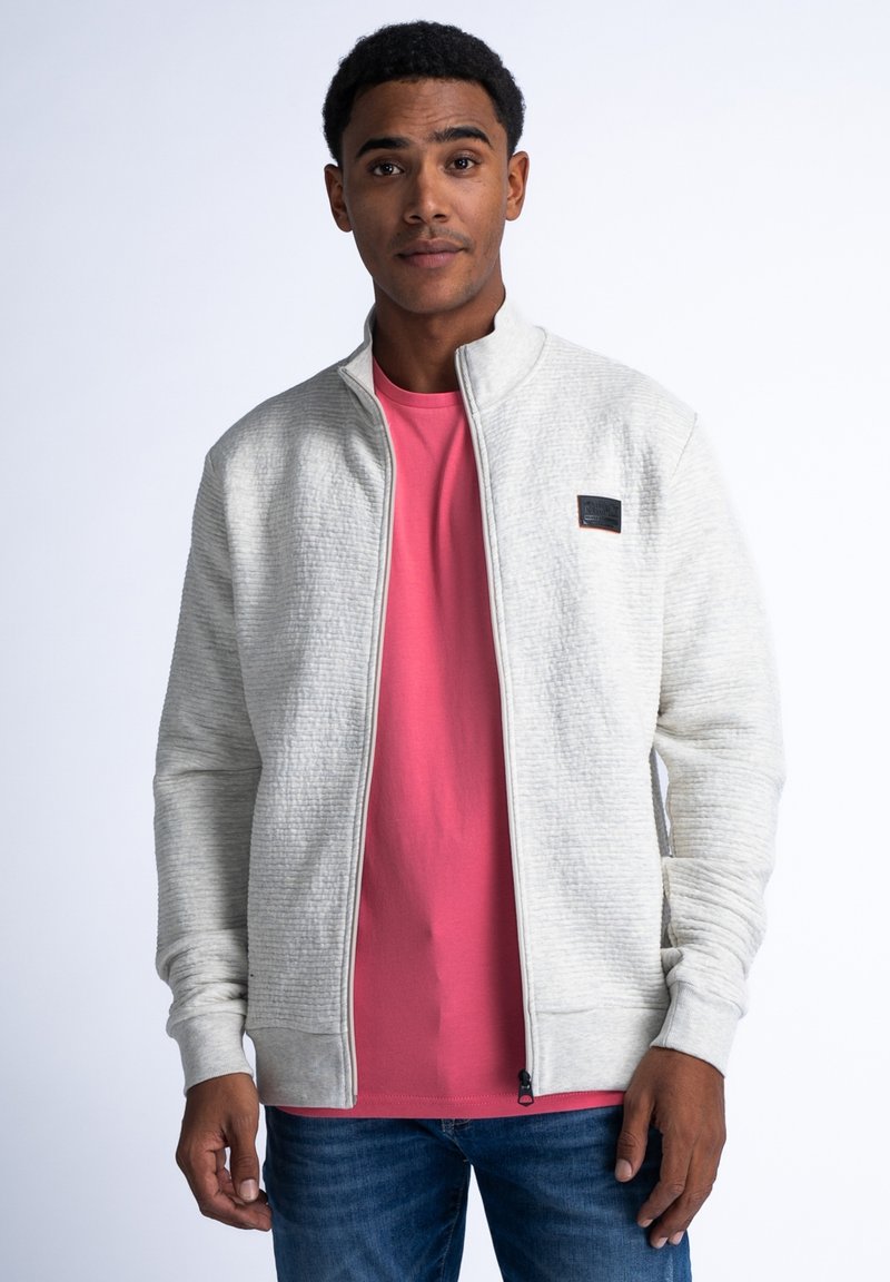 Ljusgrå zip-jacka med texturerad tyg, hög krage och ribbade mudd, bärs över en rosa skjorta. Enkel logotypdetalj på bröstet.