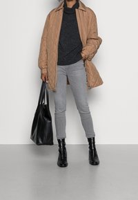 Person in einer beigefarbenen gesteppte Jacke, dunklem Pullover, grauen Jeans und schwarzen Stiefeln mit Reißverschluss vorne, hält eine große schwarze Ledertasche.