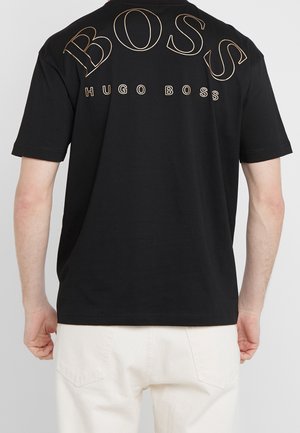 Osoba nosząca czarną koszulkę z krótkim rękawem z dużym obrysowanym tekstem "BOSS" i mniejszym tekstem "HUGO BOSS" na plecach, w połączeniu z białymi spodniami.
