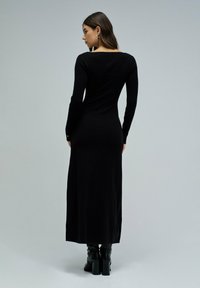 Schwarzes, langärmliges Strickkleid mit taillierter Passform, Rundhalsausschnitt und Midikleid-Länge. Getragen mit schwarzen Stiefeln mit Absätzen.