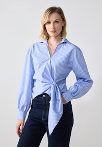 Lyseblå stripete skjorte med lange ermer, krage og knute-detalj foran, sammen med mørke jeans. Glatt stoff med en avslappet passform.