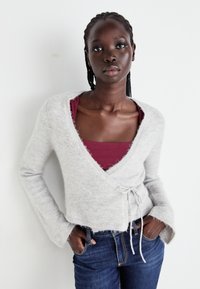 Helles graues Wickel-Strickjäckchen aus weichem, fuzzy Material mit weit geschnittenen Ärmeln, V-Ausschnitt und Bindeband. Getragen über einem burgunderfarbenen Tanktop, kombiniert mit Jeans.