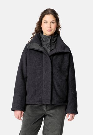 Frau trägt eine schwarze Fleecejacke über einer schwarzen, gesteppte Weste mit hohem Kragen, steht vor einem einfarbigen hellen Hintergrund.