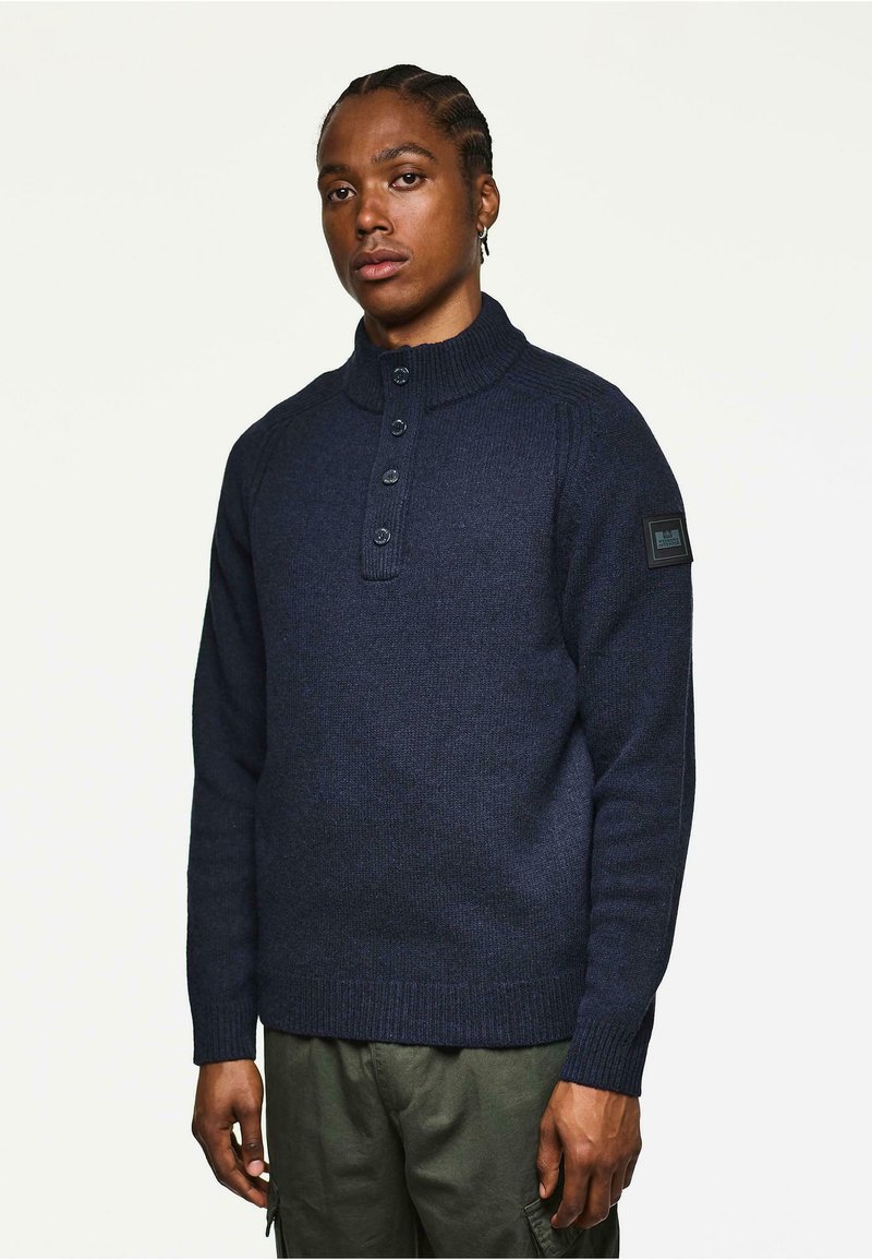 Ung mand med cornrows iført en navy blå knap-off sweater og olivengrønne cargo bukser, der står foran en ensfarvet lys baggrund.