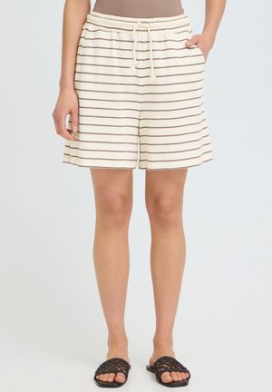 Kvinde iført cremede shorts med tynde brune vandrette striber, elastisk talje, sidelommer og sorte vævede slip-on sandaler.