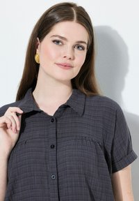 Camisa gris de botones con un patrón texturizado, mangas cortas y un cuello clásico. Presenta botones negros y un detalle fruncido en el frente.