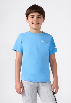 Garçon souriant avec des cheveux bruns courts portant un t-shirt Champion bleu clair et un pantalon de survêtement gris sur un fond blanc uni.