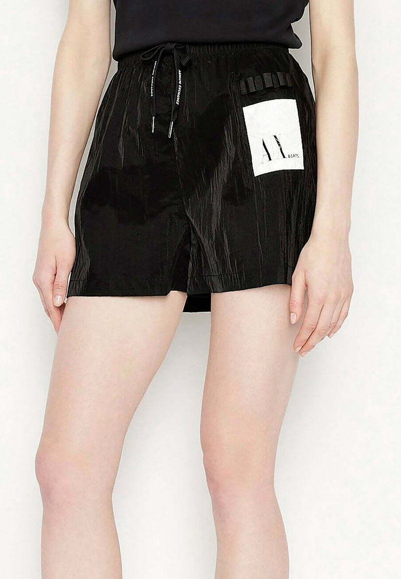 Gros plan d’une personne portant un short noir avec un patch rectangulaire blanc sur le côté droit et une taille avec cordon de serrage.