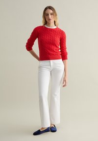 Maglione rosso a maglia rasata con maniche lunghe, pantaloni bianchi a campana e pantofole blu navy su uno sfondo neutro.