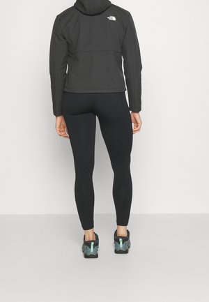 Persoon gekleed in een zwart North Face-jas met capuchon, zwarte legging en blauwgrijze sportschoenen, staand tegen een effen lichte achtergrond.