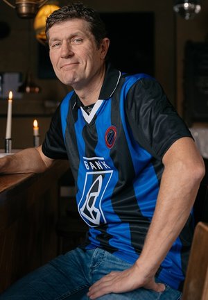 Blauw-zwart gestreept sportshirt met kraag, met een logo en de tekst "BANK" aan de voorkant. Gecombineerd met een blauwe spijkerbroek.