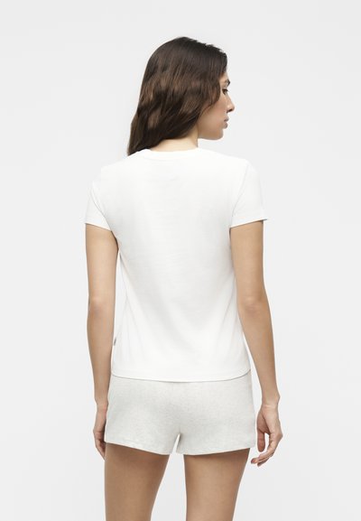 Γυναίκα με μακριά καστανά μαλλιά, που φοράει απλό λευκό κοντομάνικο μπλουζάκι και ανοιχτό γκρι σορτς, στέκεται με την πλάτη σε λευκό φόντο.