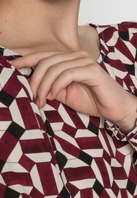 Haut à manches longues présentant un motif géométrique en bordeaux, beige et noir. Tissu doux avec une texture lisse, coupe ajustée.