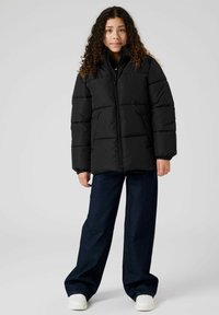 Chaqueta de plumas negra con cuello alto, que presenta un cierre de cremallera frontal, diseño acolchado y puños elásticos, combinada con jeans de denim de pierna ancha y zapatillas blancas.