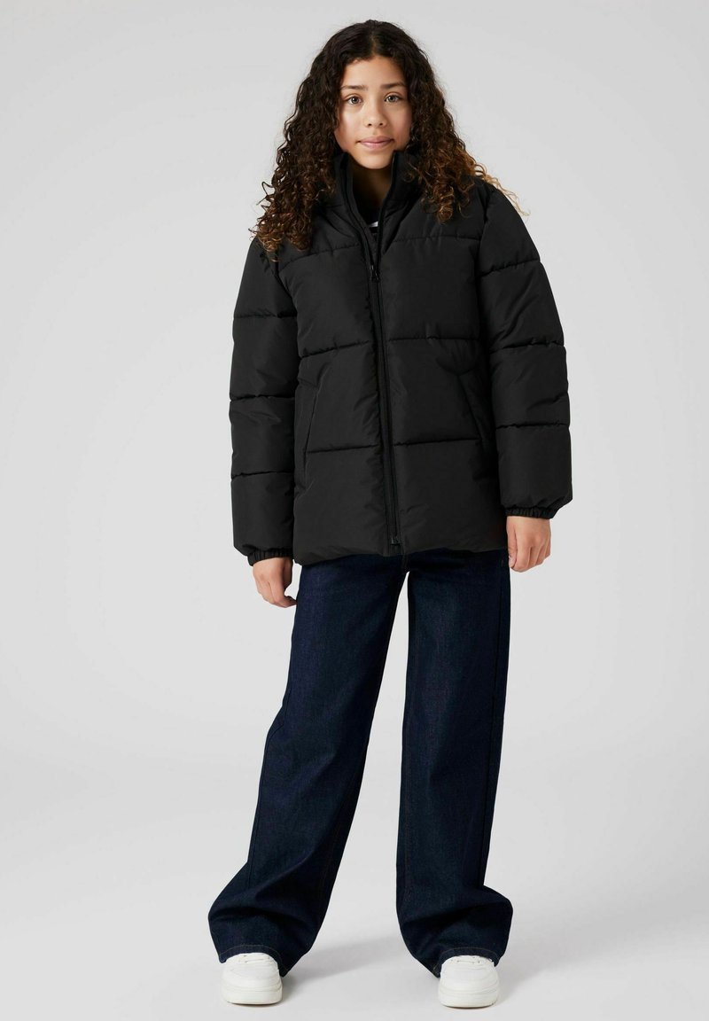 Chaqueta de plumas negra con cuello alto, que presenta un cierre de cremallera frontal, diseño acolchado y puños elásticos, combinada con jeans de denim de pierna ancha y zapatillas blancas.