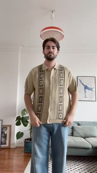 Camicia button-up beige con un motivo geometrico scuro sul davanti. Maniche corte, vestibilità rilassata, abbinata a pantaloni azzurri chiari.